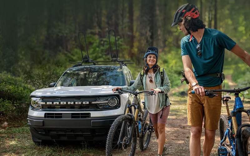 A couple about to go for a bike ride in a wooded area with a white Ford Bronco parked in the background